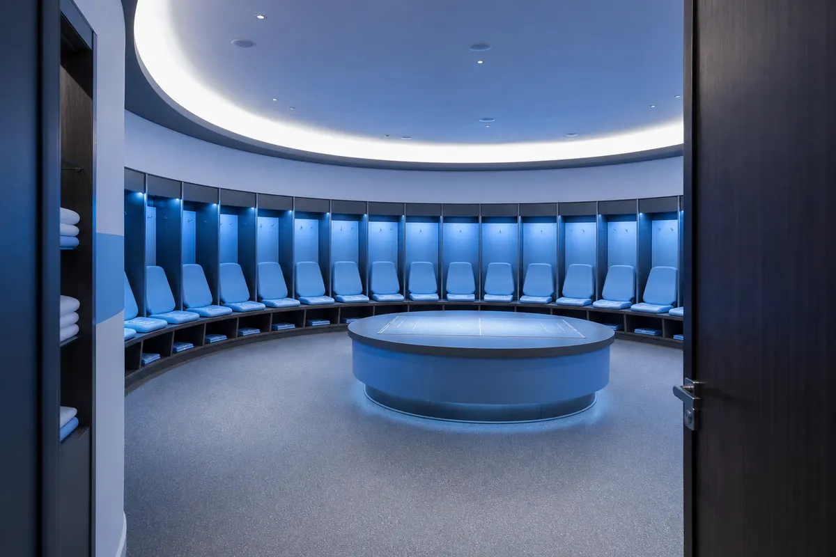 Modern football dressing room with pale blue seats, lockers and a central tactics table, illustrating a typical Manchester City stadium tour stop
