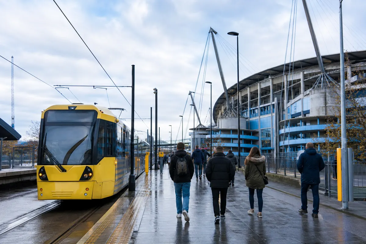 Visitors arriving by yellow city tram beside a large Manchester football stadium on a wet day